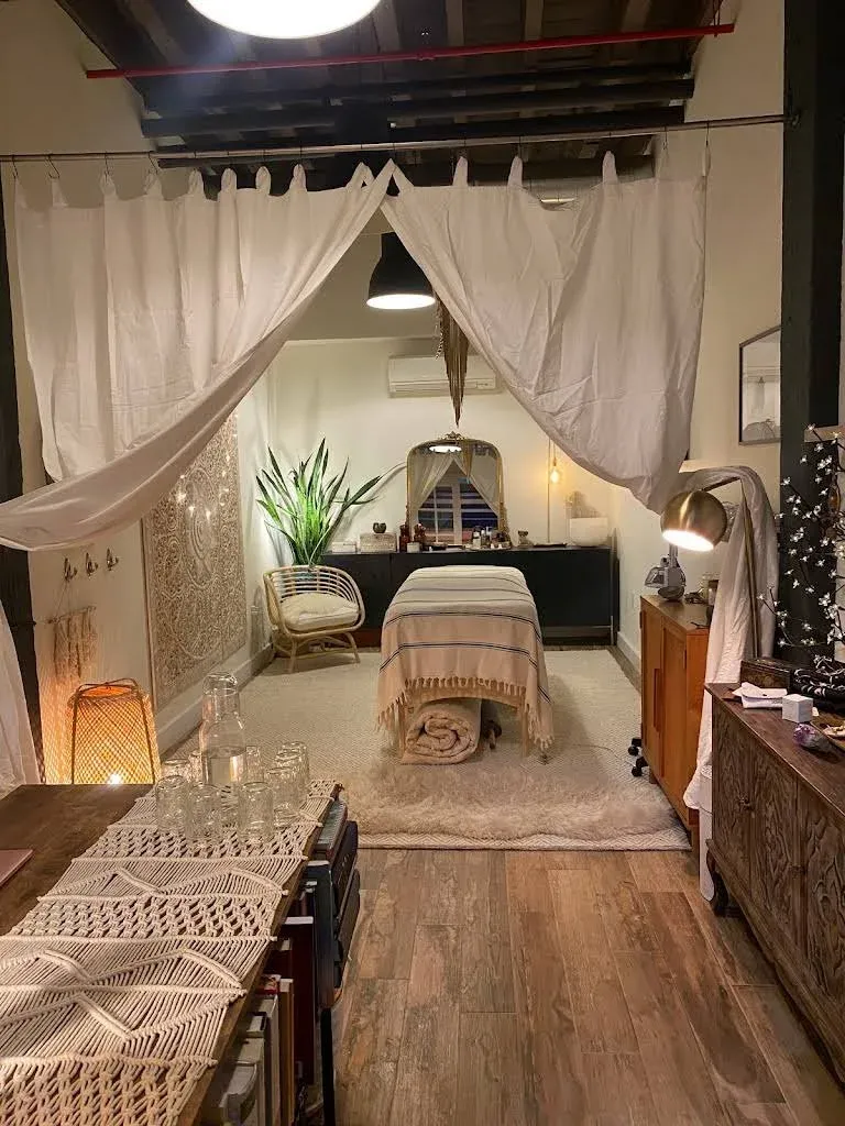 Cozy massage room with a massage table, soft lighting, plants, white curtains, wooden floors, and decorative accents like a woven lamp and a tapestry.