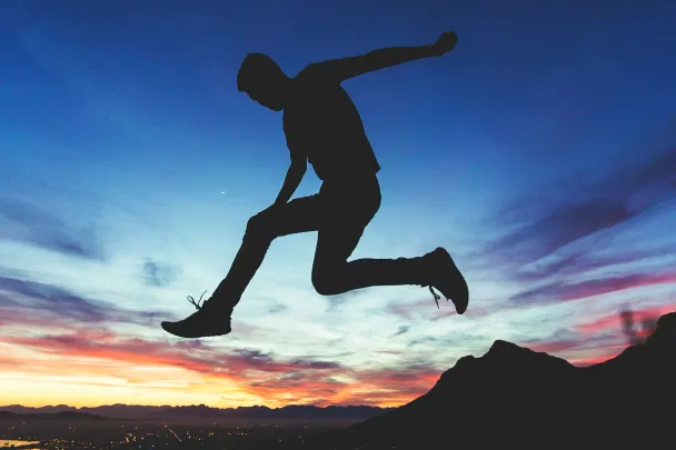 Silhouette of a person jumping against a sunset sky with clouds and mountains in the background.