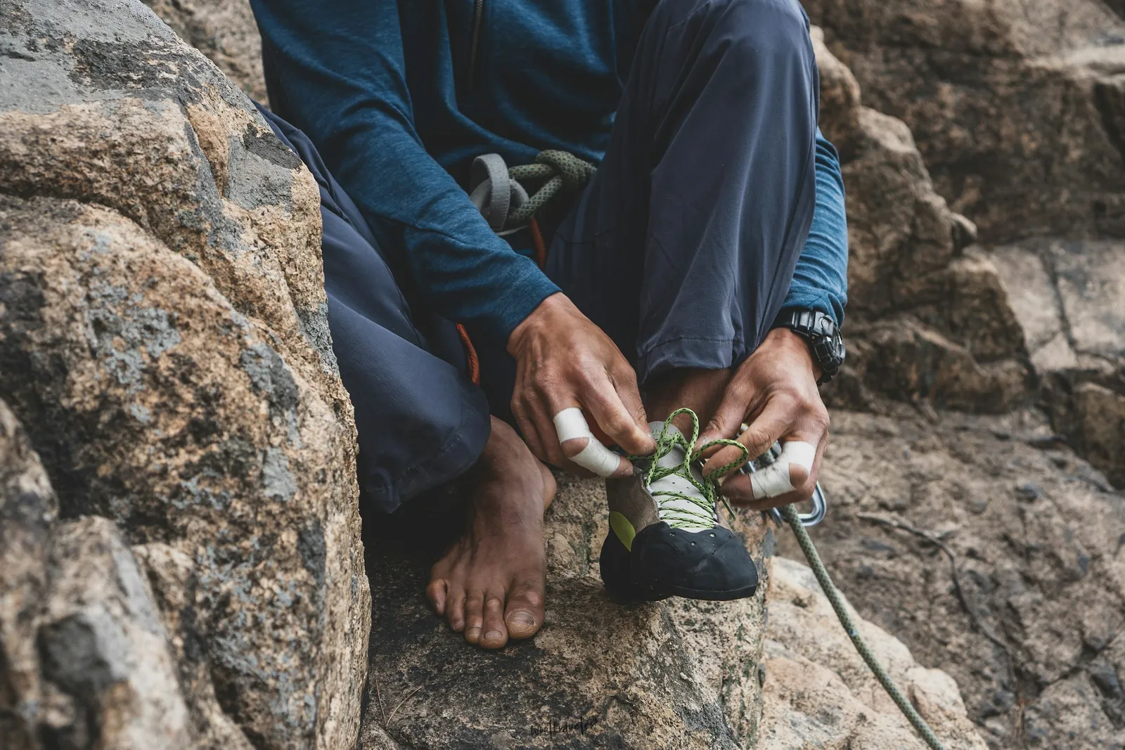 A climber with taped fingers pulling a climbing shoe onto one bare foot, sitting on rock