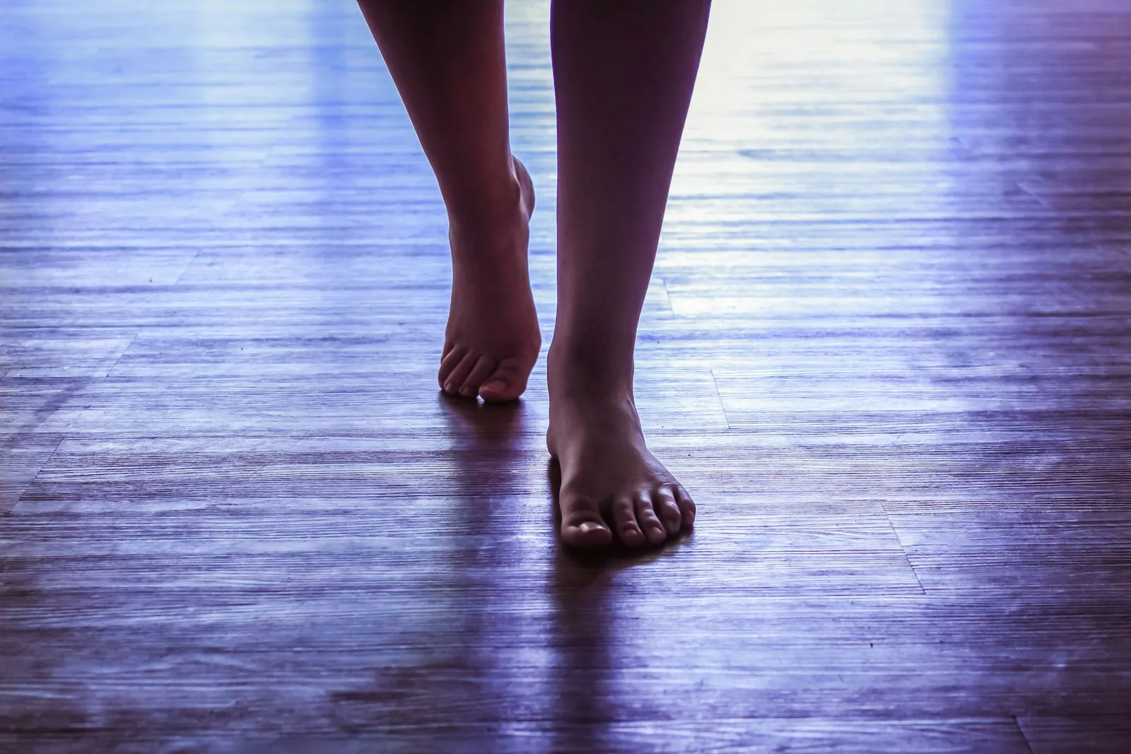 Bare feet standing on a wooden floor in low light, one heel lifted
