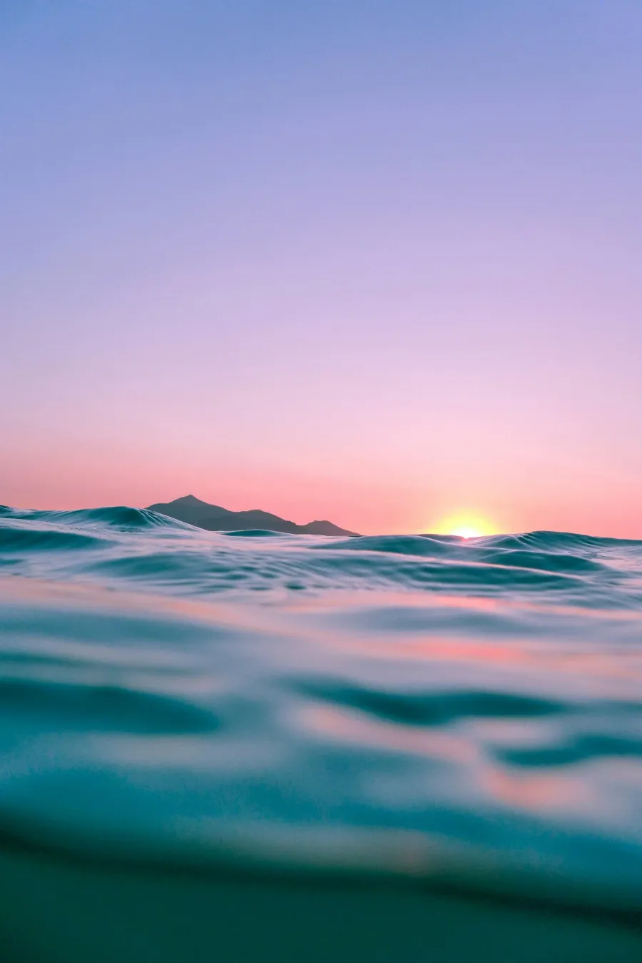 Ocean waves at sunrise or sunset with distant mountains and colorful sky.